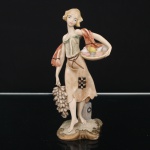 Escultura decorativa em material resina moldada, representando figura feminina trajando vestes estilizadas, portando cesto com conchas marinhas e cacho de uvas. Base moldada com elementos ornamentais em relevo e policromia aplicada. Medidas: Alt.: 20,5 x Comp.: 11 x Larg.: 8 cm.