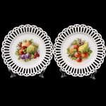 Dois(02)  pratos decorativos em porcelana, com borda vazada recortada em padrão rendilhado. Centro ornamentado por composições policromadas representando frutos, incluindo maçãs, peras, ameixas, melão e frutos silvestres. Decoração aplicada por transferência e esmaltação sobre fundo branco. Medida: Diam.: 19 cm.