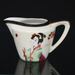 Leiteira em porcelana japonesa, de corpo ligeiramente obcônico e bico vertedor ondulado, com alça estilizada integrada. Decoração policromada representando figura feminina em trajes tradicionais japoneses, acompanhada por ramos de folhas estilizadas. Borda realçada por friso em tonalidade escura e contorno com filete dourado. Base marcada com inscrição Made in Japan. Medidas: Alt.: 7,5 x Comp.: 13 x Larg.: 8,5 cm.