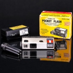 Câmera fotográfica compacta Kodak Pocket Instamatic 20, produzida para utilização de filme cartucho formato 110, com corpo retangular em metal e material plástico, visor frontal e disparador superior. Acompanha flash eletrônico repetitivo Acme-Lite Model 1060, destinado a câmeras de bolso, com respectiva caixa original ilustrada, além de suporte empunhadura destacável. Conjunto representativo da fotografia amadora portátil das décadas de 1970 e 1980. Apresenta sinais de uso. Não foi testada, sem garantias de funcionamento.Medidas máquina: Alt.:3 x Comp.: 11,5 x Larg.: 5,5 cm.Medidas flash: Alt.: 4 x Comp.: 8,5 x Larg.: 6,5 cm.