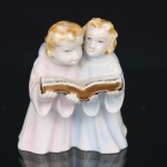 Grupo escultórico em porcelana representando dois anjos infantis em atitude de canto, segurando livro, trajando túnicas em tons rosa e azul, e detalhes dourados. Modelagem naturalista com policromia aplicada. Medidas: Alt.: 10 x Comp.: 8,5 x Larg.: 6 cm.