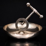 WOLFF. Quebra-nozes de mesa em metal prateado, composto por prato circular de apoio e mecanismo de compressão por rosqueamento. Estrutura formada por suporte helicoidal aberto, haste roscada central e manípulo transversal com terminais esféricos. Destinado à quebra de nozes, castanhas e frutos secos de casca rígida. Marca da manufatura no verso. Medidas: Alt.: 18 x Diam.: 18 cm.