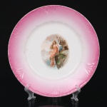 Prato de coleção em porcelana francesa Jean Pouyat, Limoges, com aba em esmalte rosa degradê moldada com reservas em relevo radial e filete dourado na borda. Ao centro, apresenta cena galante de inspiração simbolista, representando figura feminina alada em paisagem rochosa, executada por decalcomania policromada. Produção francesa do início do século XX, vinculada ao estilo Art Nouveau, com temática alegórica e tratamento pictórico característico das porcelanas decorativas de Limoges. Marcada no vers. Medida: Diam.: 22,5 cm.