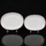 Porcelana Renner, Porto Alegre - RS(Fundada em 1937). Duas(02) travessas oblongas em porcelana nacional da década de 1960/70; esmaltação na cor branca, borda ornada por cercadura em meandro contínuo, de tonalidade cinza, e sucessão de quadrados duplos em douração aplicada. Marca da manufatura no verso. Medidas: Comp.: 34 x Larg.: 23 cm. ; Medidas: Comp.: 29,5 x Larg.:  20 cm.