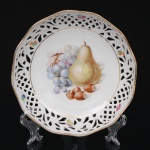 Pequena fruteira de borda vazada em porcelana alemã Schumann Arzberg, decoração policroma representando natureza-morta com pera, cacho de uvas e avelãs ao centro, aba recortada com trabalho rendilhado vazado e reservas com pequenas frutas, arrematada por filetes dourados. Marca da manufatura no verso. Medida: Diam.: 19 cm.