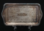 Bandeja retangular em metal espessado a prata, manufatura Bellini, com borda moldurada por friso perlado e elementos de concheados nos cantos. Campo central gravado com composição ornamental de arabescos, cartelas e folhagens, contendo dedicatória nominativa Lucy e inscrição com citação atribuída a Fernando Pessoa, datada de 29.02.1980. Necessita refazer banho de prata. Medidas: Comp.: 36,5 x Larg.: 20 cm.