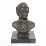Busto de Wolfgang Amadeus Mozart em pétit bronze, representado com traje de época, identificado na base pela inscrição MOZART, apoiado sobre plinto retangular em mármore negro. Medidas: Alt.: 16 x Comp.: 9,5 x Larg.: 9,5 cm.