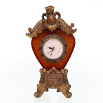 Relógio de mesa decorativo em resina moldada, corpo em formato de cartela recortada com acabamento marmorizado em tons âmbar e realces dourados, mostrador circular com numeração romana, cercadura floral estilizada, encimado por ornatos fitomórficos e botão floral, apoiado sobre base recortada com elementos rocaille. Marca Dimontti Italy Style. Não está funcionando. Medidas: Alt.: 25,5 x Comp.: 14,5 x Larg.: 9 cm.