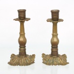 Par de castiçais em bronze, fuste torneado de seção balaustrada com elementos fitomórficos em relevo, bobeches em forma de folha estilizada e bases recortadas decoradas por folhagens e volutas. Medidas: Alt.: 18 x Diam.: 9,5 cm.