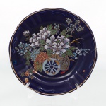 Prato decorativo em porcelana vitrificada de fundo azul-cobalto, borda recortada com filete dourado, ornamentado ao centro com composição floral policromada em cesta sobre rodas, executada em estampagem e douração. Medida: Diam.: 21 cm.