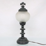 Abajur de mesa em metal fundido patinado, com haste torneada e ornamentação de inspiração neobarroca, montado com globo esférico em vidro fosco gravado com motivos estrelados. Eletrificado. Apresenta oxidação, desgaste da pátina e sinais de uso. Medidas: Alt.: 41 x Diam.: 15,5 cm.