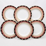 Porcelana, Steatita e Magnésia S/A, Campo Largo - PR(Fundada em 1954). Conjunto de pratos rasos em porcelana nacional da década de 1960/70; decoração em composição concêntrica, borda externa em esmalte preto ornada por reservas circulares em branco, intercaladas de maneira ritmada, contornada por filetes dourados. Faixa subsequente em esmalte rosa com sequência de grinaldas e elementos fitomorfos estilizados aplicados em douração. Cercadura interna em fundo nacarado com motivos radiais e palmetas estilizadas em ouro brunido. Calha delimitada por friso contínuo em douração rendilhada, formando sequência de volutas e elementos anelares. Contorno ondulado com acabamento dourado aplicado à borda. Composto de seis(06) unidades. Medida: Diam.: 24,5 cm.