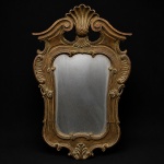 Espelho de parede em madeira entalhada e dourada. Moldura recortada, vazada e desenvolvida por curvas e contracurvas. Frontão centrado por concha em relevo, ladeada por volutas espiraladas e reservas vazadas. Decoração composta por elementos concheados, enrolamentos, folhas de acanto estilizadas, cartelas recortadas e motivos fitomorfos distribuídos ao longo da estrutura. Laterais com reservas em relevo de nervuras radiais, remetendo a conchas abertas. Moldura interna acompanhando o contorno sinuoso da peça, com terminais volutados nos segmentos superior e inferior. Base ornada por folha de acanto centralizada e enrolamentos afrontados integrados ao desenho da moldura. Verso com fechamento em madeira e papel de proteção remanescente fixado manualmente. Medidas: Alt.: 64,5 x Comp.: 40 cm.