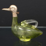 Galheta em cristal soprado, tonalidade chartreuse, executada na forma de ave, tampa representando a cabeça do animal, confeccionada em metal prateado, com movimento basculante para despejo do líquido, alças aplicadas na superfíei representando as asas. Medidas: Alt.: 16,5 x Comp.: 19 x Larg.: 9 cm.