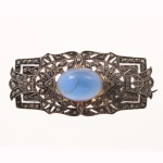 Broche confeccionado em prata, com trabalho vazado de inspiração Art Déco, ornamentado por marcassitas em cravação de grão(com algumas perdas) e cabochão oval de vidro opalescente azul em engaste fechado. Apresenta composição geométrica e rendilhada característica da joalheria das primeiras décadas do século XX, com fecho de alfinete articulado(necessita ajuste). Medidas: Comp.: 5 x Larg.: 2,5 cm. Peso total: 11.7 gramas.