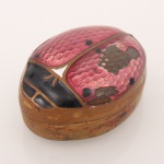 Caixa oval confeccionada em metal dourada, com tampa articulada ornamentada por esmaltação alveolada policroma em tons de rosa, preto e branco, formando composição representando joaninha. Apresenta desgastes, oxidações e perdas na camada de esmalte. Medidas: Alt.: 1,5 x Comp.: 2,8 x Larg.: 3,5 cm.