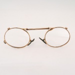 Óculos tipo pince-nez, com armação oval em metal dourado, ponte central dotada de mola helicoidal de pressão, lentes incolores e apoio nasal ajustável. Trabalho do final do século XIX ou início do século XX. Acompanha estojo original revestido internamente em veludo roxo. Apresenta desgastes, oxidações e perdas compatíveis com a idade e o uso. Medidas: Comp.: 10 x Larg.: 3,5 cm.