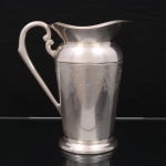EBERLE. Elegante jarra confeccionada em metal espessurado à prata, bojo balaústre ricamente lavrada com elementos vegetalistas, assente sobre base circular repuxada em degraus, asa lateral em volutas, boda ondulada e revirada para fora, interior dotado de barra retentora de gelo. Marca da manufatura ao fundo. Med.: Alt. 22 x Comp. 20 x Larg. 13 cm. Apresenta marcas de uso.