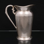 EBERLE. Elegante jarra confeccionada em metal espessurado à prata, bojo balaústre ricamente lavrada com elementos vegetalistas, assente sobre base circular repuxada em degraus, asa lateral em volutas, boda ondulada e revirada para fora, interior dotado de barra retentora de gelo. Marca da manufatura ao fundo. Med.: Alt. 22 x Comp. 20 x Larg. 13 cm. Apresenta marcas de uso.