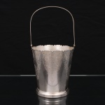 REGIA PRATA 90. Balde para gelo em metal prateado, de corpo obcônico, assente sobre base circular e provido de alça móvel. Borda recortada em ondulações e decoração gravada por toda a superfície com motivos de flores, volutas e elementos fitomórficos dispostos em reservas repetidas. Marca da manufatura ao fundo. Apresenta sinais de uso e pequenos desgastes. Medidas. Alt.: 26 x Diam.: 12 cm.