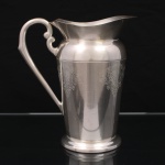 EBERLE. Elegante jarra confeccionada em metal espessurado à prata, bojo balaústre ricamente lavrada com elementos vegetalistas, assente sobre base circular repuxada em degraus, asa lateral em volutas, boda ondulada e revirada para fora, interior dotado de barra retentora de gelo. Marca da manufatura ao fundo. Med.: Alt. 22 x Comp. 20 x Larg. 13 cm. Apresenta marcas de uso.