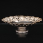 REGIA PRATA 90. Fruteira em metal prateado, de formato circular e interior côncavo, assente sobre base alteada, borda recortada e vazada em repetição de cartelas rendilhadas, ornamentadas por flores estilizadas, folhagens e volutas gravadas e recortadas. Apresenta sinais de uso e leves desgastes do banho de prata. Marca da manufatura no verso. Medidas: Alt.: 7 x Diam.: 23 cm.