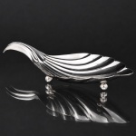 Elegante petisqueira confeccionada em metal espessurado à prata, executada na forma de concha, interior côncavo repuxado com sulcos ondulados, alça lateral curvada para dentro, assente sobre três pés esféricos. Med.: Alt. 4 x Comp. 20 x Larg. 9,5 cm.