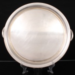 Elegante bandeja circular confeccionada em metal espessurado à prata, centro ligeiramente rebaixado, borda de contorno elevado, base alargada, alças laterais em arco terminado em volutas. Apresenta marcas de uso, desgastes no banho de prata, e pequenos trincados. Demanda restauro. Medidas: Alt.: 3,5 x Diam.: 44 cm.