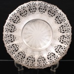 REGIA PRATA 90. Fruteira circular confeccionada em metal prateado, com borda recortada em ondulações sucessivas e ornamentação rendilhada obtida por vazados de motivos fitomórficos entrelaçados, gravados e perfurados, formando composição repetitiva de arabescos e volutas. Cavidade central rebaixada, ornada por desenho radial gravado composto por folhas estilizadas dispostas em torno de núcleo circular. Assentada sobre três pés aplicados na face inferior. Peça destinada ao serviço e apresentação de frutas, doces ou confeitos. Apresenta desgastes e marcas de uso compatíveis com o tempo. Marca da manufatura no verso. Medida: Alt.: 6,5 x Diam.: 37 cm.