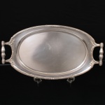 WOLFF. Elegante bandeja oblonga confeccionada em metal espessurado à prata, centro oval ligeiramente rebaixado, borda contornada por fileira de frisos intercalados por fitas cruzadas e flores, alças laterais fusiformes com terminações cobertas por folhagens relevadas. Marca da manufatura puncionada no verso. Med.: Alt. 5 x Comp. 54 x Larg. 30 cm. Apresenta leves marcas de uso.