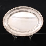 FRACALANZA. Elegante travessa confeccionada em metal espessurado à prata, formato oval, centro ligeiramente rebaixado, borda relevada com fileira de frisos intercaladas por fitas cruzadas, ornato obtido através da técnica do cinzelado. Marca da manufatura no verso. Apresenta marcas de uso. Medida: Comp.: x Larg.:  cm.