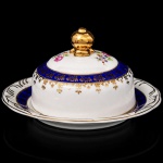 Porcelana Schmidt - Brasil(Fundada em 1945). Exuberante manteigueira em porcelana nacional da década de 1950/60, executada ao estilo francês do período Luís XV; esmaltagem vítrea na cor branca, circundada por faixas de tonalidade azul cobalto, profusamente realçada com frisos e faixa com repetição de motivos fitomorfos em douradura; reservas florais compostas por rosas, violetas e pequenas flores campestres, distribuídas na tampa, puxador modelado em forma de fruto sobre laçarote. Marca da manufatura ao fundo. Medidas: Alt.: 8,5 x Diam.: 15,5 cm.