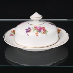 Porcelana Real, Mauá - SP(Fundada em 1943 por Fritz Erwin Schmidt). Manteigueira em porcelana nacional da década de 1950/60; esmaltação branca, decorada com padrão floral em estampagem policromada, distribuídas em arranjos na tampa, e com flores soltas ao redor do presentoir; contornos realçados por filete em douradura; borda recortada em arcos, tampa abaulada encimada por puxador com feitio de pião. Marca da manufatura ao fundo. Medidas: Alt.: 7 x Diam.: 15 cm.