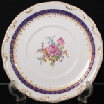 Porcelana Schmidt - Brasil(Fundada em 1945). Exuberante prato para serviço de sobremesa em porcelana nacional da década de 1950/60, executada ao estilo francês do período Luís XV; esmaltagem vítrea na cor branca, circundada por faixas de tonalidade azul cobalto, profusamente realçada com frisos e faixa com repetição de motivos fitomorfos em douradura; reserva floral compostas por rosas, violetas e pequenas flores campestres ao centro, orla relevada com aplicações em relevo ao longo da aba. Marca da manufatura ao fundo. Medidas: Diam.: 28 cm.