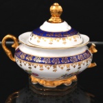 Porcelana Schmidt - Brasil(Fundada em 1945). Exuberante açucareiro em porcelana nacional da década de 1950/60, executada ao estilo francês do período Luís XV; esmaltagem vítrea na cor branca, circundada por faixas de tonalidade azul cobalto, profusamente realçada com frisos e faixa com repetição de motivos fitomorfos em douradura; reservas florais compostas por rosas, violetas e pequenas flores campestres, distribuídas no bojo, tampa campaniforme encimada por puxador modelado em forma de fruto sobre laçarote, alças laterais em voluta, base circular recortada. Marca da manufatura ao fundo. Medidas: Alt.: 11 x Comp.: 14 x Larg.: 11,5 cm(apresenta um trincado no bojo e falta uma alça, vide foto).
