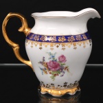 Porcelana Schmidt - Brasil(Fundada em 1945). Exuberante leiteira em porcelana nacional da década de 1950/60, executado ao estilo francês do período Luís XV; esmaltagem vítrea na cor branca, faixas de tonalidade azul cobalto, profusamente realçada com frisos, faixa com repetição de motivos fitomorfos em padrão seriado; reservas florais compostas por rosas, violetas e pequenas flores campestres, distribuídas no bojo; bico vertedor ondulado e revirado para fora, asa lateral em grande voluta, base circular recortada. Marca da manufatura ao fundo. Medidas: Alt.: 12 x Comp.: 14,5 x Larg.: 10,5 cm.