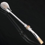 Bomba para chimarrão confeccionada em metal prateado, haste tubular e passador relevado com flores e folhagens, patilha oval perfurada, boquilha achatada e levemente curvada. Medida: Comp.: 20 cm..