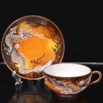 Trio para chá em porcelana japonesa, dita "casca de ovo", bojo hemisférico decorado com tonalidades de âmbar, ocre e castanho, e representação de dragão oriental modelado em relevo e aplicado sobre a superfície, com escamas, garras, bigodes e crista destacados, complementado por faixas sinuosas e elementos lineares que remetem à composição de nuvens e movimento, distribuídos sobre fundo em esmaltação graduada, decoração replicada no pires e no prato. Bordas e alça arrematados em douradura. Marcas de manufatura na base, Made in Japan. Medidas: Xícara: Alt.: 4,5 x Comp.: 11 x Larg.: 9 cm; Pires: 13,5 cm; Prato: Diam.: 17 cm.
