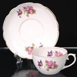 Porcelana Real, Mauá - SP(Fundada em 1943 por Fritz Erwin Schmidt). Xícara para chá em porcelana nacional da década de 1950/60; esmaltação branca, bojo hemisférico e alça lateral em voluta, decorada com padrão floral em estampagem policromada, contornos realçados por filete em douradura; pires com borda recortada em arcos. Marca da manufatura ao fundo. Medidas: Xícara: Alt.: 5 x Comp.: 10,5 x Larg.: 8,5 cm; Medidas: Pires: Diam.: 14,5 cm.