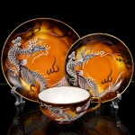 Trio para chá em porcelana japonesa, dita "casca de ovo", bojo hemisférico decorado com tonalidades de âmbar, ocre e castanho, e representação de dragão oriental modelado em relevo e aplicado sobre a superfície, com escamas, garras, bigodes e crista destacados, complementado por faixas sinuosas e elementos lineares que remetem à composição de nuvens e movimento, distribuídos sobre fundo em esmaltação graduada, decoração replicada no pires e no prato. Bordas e alça arrematados em douradura. Marcas de manufatura na base, Made in Japan. Medidas: Xícara: Alt.: 4,5 x Comp.: 11 x Larg.: 9 cm; Pires: 13,5 cm; Prato: Diam.: 17 cm.