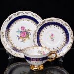 Porcelana Schmidt, Santa Catarina - Brasil(Fundada em 1945). Exuberante trio para chá em porcelana nacional da década de 1950/60, executado ao estilo francês do período Luís XV; esmaltagem vítrea na cor branca, faixas de tonalidade azul cobalto, profusamente realçada com frisos, faixa com repetição de motivos fitomorfos em padrão seriado; reservas florais compostas por rosas, violetas e pequenas flores campestres, distribuídas ao centro do prato e do pires, bem como no interior da xícara complementam a composição, bojo hemisférico assente sobre base alteada, asa lateral em voluta. Marca da manufatura ao fundo. Medidas: Chávena: Alt.: 5,5 x Comp.: 11,5 x Larg.: 9,5 cm.