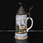 Tankard de reservista (Reservistenkrug) em porcelana, com tampa articulada em estanho e terminal escultórico representando águia. Corpo cilíndrico sobre base escalonada, decorado em policromia com brasão bávaro encimado por coroa, bandeiras, militares uniformizados, dirigível, aeronaves, âncora, peças de artilharia e inscrições nominativas em alemão. Tampa com cercadura gravada e acionador vazado de motivo fitomórfico. A decoração faz referência ao serviço militar e à aviação bávara, incluindo relação de integrantes da unidade e inscrições alusivas ao período de serviço. Alemanha, início do século XX. A inscrição "Luftschiffer" (aeronauta/aviador) torna a peça particularmente associada às unidades alemãs de aviação militar anteriores à Primeira Guerra Mundial. Medidas: Alt.: 27 x Comp.: 14 x Larg.: 11 cm.
