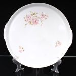 Travessa circular de serviço, em porcelana, com duas pequenas pegas laterais integradas à aba. Decorada com composição floral de rosas em tonalidade rosa e folhagens distribuídas junto à borda. Destinada ao serviço de mesa para doces, bolos e petit fours. Marcada na base Medaillon Porcelana Renner, Porto Alegre. Medida: Diam.:
