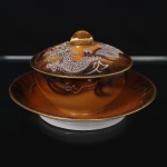 Manteigueira em porcelana japonesa, dita "casca de ovo", bojo hemisférico decorado com tonalidades de âmbar, ocre e castanho, e representação de dragão oriental modelado em relevo e aplicado sobre a superfície, com escamas, garras, bigodes e crista destacados, complementado por faixas sinuosas e elementos lineares que remetem à composição de nuvens e movimento, distribuídos sobre fundo em esmaltação graduada, tampa abaulada encimada por puxador em dupla folhagem. Bordas e pega arrematados em douradura. Marcas de manufatura na base, Made in Japan. Medidas: Alt.: 7,5 x Diam.: 13,5 cm.
