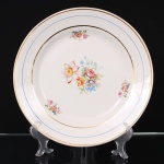 Prato em faiança nacional da década de 1940/50, esmaltagem de tom marfim, decorado com composição floral policroma ao centro e reservas florais distribuídas na aba. Borda contornada por frisos dourados e filete azul. Marca da manufatura na base. Medida: Diam.: 28 cm.