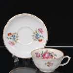 C.T. Tielsch, Walbrzych (antiga Altwasser) - Polônia. Belísima xícara para chá em porcelana polonesa da década de 1390/40; esmaltagem vítrea na cor branca, decorada em policromia com composição floral constituídas por rosas, tulipas e flores campestres, distribuídas em reservas no interior da xícara, no bojo e no pires, aplicada em estampagem; bordas realçadas por filetes dourados. Marca da manufatura no verso. Medidas: Xícara: Alt.: 5 x Comp: 12 x Larg.: 9,5 cm. ; Pires: Diam.: 15 cm.