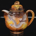 Leiteira em porcelana japonesa, dita "casca de ovo", bojo oval decorado com tonalidades de âmbar, ocre e castanho, e representação de dragão oriental modelado em relevo e aplicado sobre a superfície, com escamas, garras, bigodes e crista destacados, complementado por faixas sinuosas e elementos lineares que remetem à composição de nuvens e movimento, distribuídos sobre fundo em esmaltação graduada, tampa abaulada encimada por puxador em dupla folhagem, bico vertedor revirado para fora, asa lateral curvada. Bordas, pega e alça arrematados em douradura. Marcas de manufatura na base, Made in Japan. Medidas: Alt.: 12 x Comp.: 13 x Larg.: 9,5 cm. Apresenta dois pequenos desgastes na douradura da borda da tampa.
