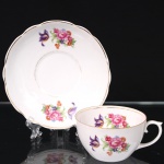 Porcelana Real, Mauá - SP(Fundada em 1943 por Fritz Erwin Schmidt). Xícara para chá em porcelana nacional da década de 1950/60; esmaltação branca, bojo hemisférico e alça lateral em voluta, decorada com padrão floral em estampagem policromada, contornos realçados por filete em douradura; pires com borda recortada em arcos. Marca da manufatura ao fundo. Medidas: Xícara: Alt.: 5 x Comp.: 10,5 x Larg.: 8,5 cm; Medidas: Pires: Diam.: 14,5 cm.