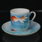 Xícara de café com respectivo pires em porcelana japonesa, decoração policroma representando peixe ornamental em tonalidades laranja sobre fundo azul degradê, com elementos vegetalistas estilizados. Marca da manufatura japonesa na face inferior, composta por escudo com monograma N sob sol nascente, acompanhada de caracteres japoneses. Apresenta fio de cabelo no corpo da xícara. Medidas: Xícara: Alt.: x Comp.: x Larg.: ; Pires: Diam.: