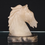 Escultura representando cabeça de cavalo, em alabastro esculpido, apoiada sobre base retangular em pedra, crina estilizada em relevo e tratamento volumétrico simplificado. Medidas: Alt.: 20 x Comp.: 16 x Larg.: 7 cm.